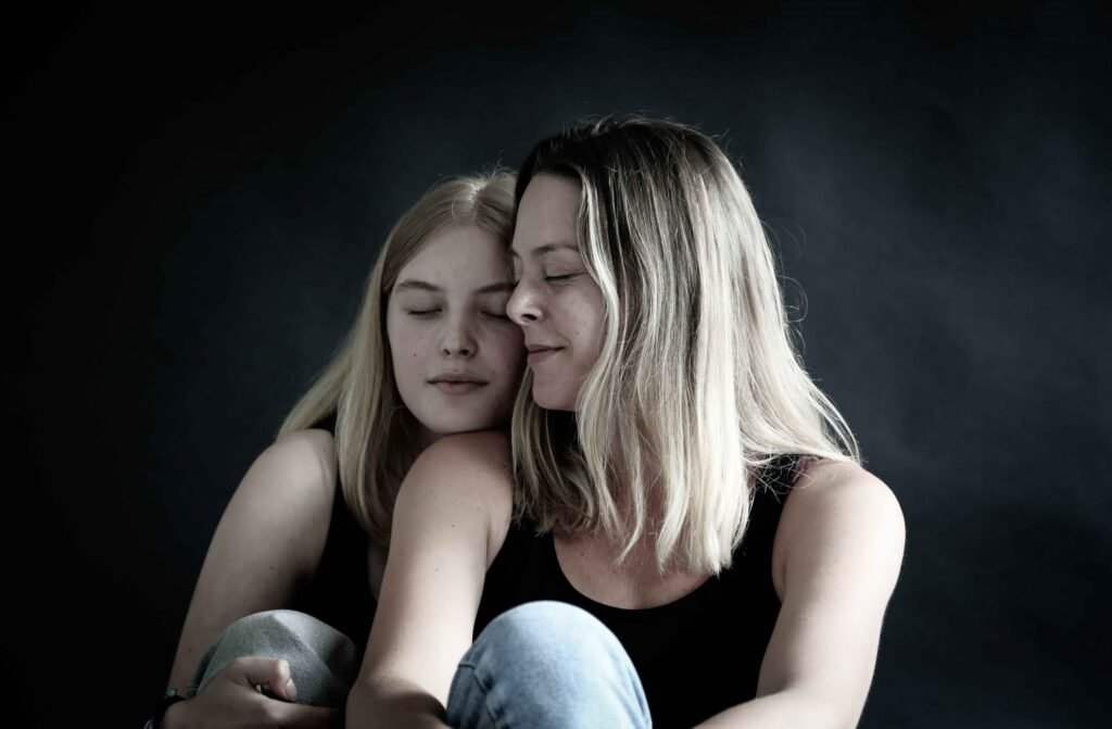 Mother and teenage daughter sitting closely with eyes closed against a dark backdrop in a serene family portrait.