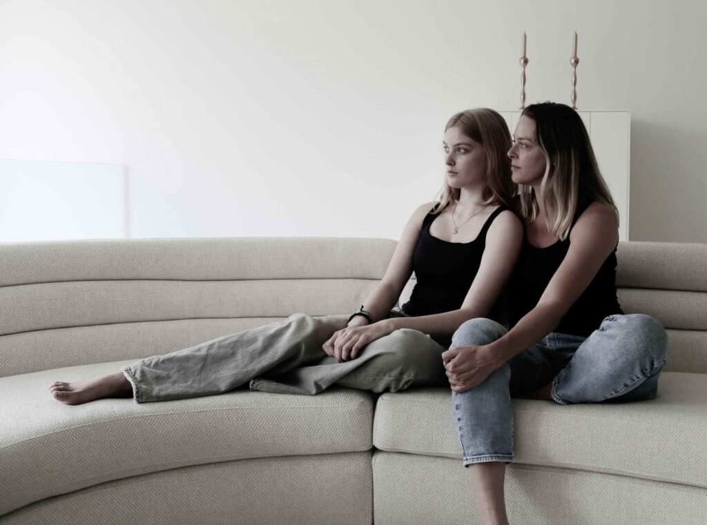 Mother and teenage daughter sitting closely on a curved beige sofa during a minimalist family photoshoot in The Hague.