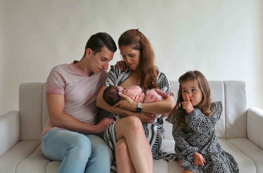 Parents with newborn and older daughter on a couch; mother feeding baby while daughter gestures “shh” in a playful moment.