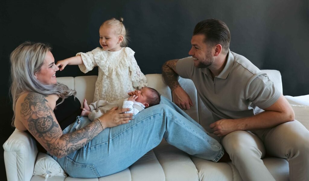 Family on a couch with mother holding newborn while toddler playfully touches her face and father looks on smiling.