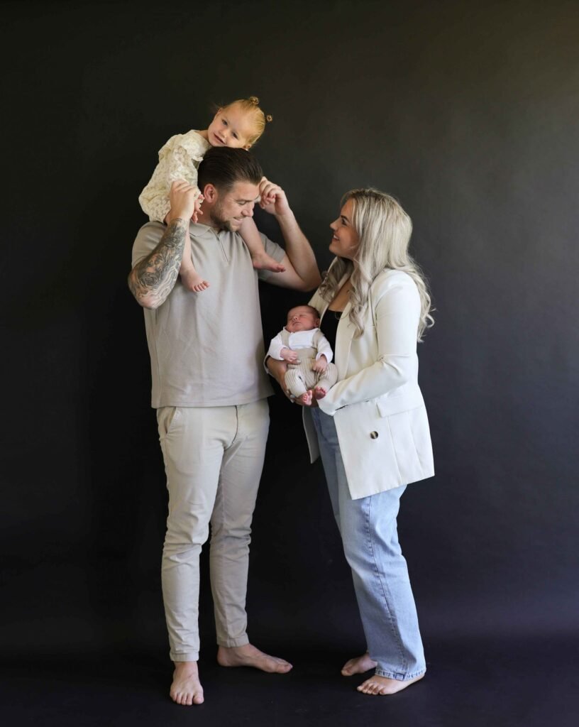 Family with toddler on father’s shoulders and newborn in mother’s arms during a studio photoshoot.