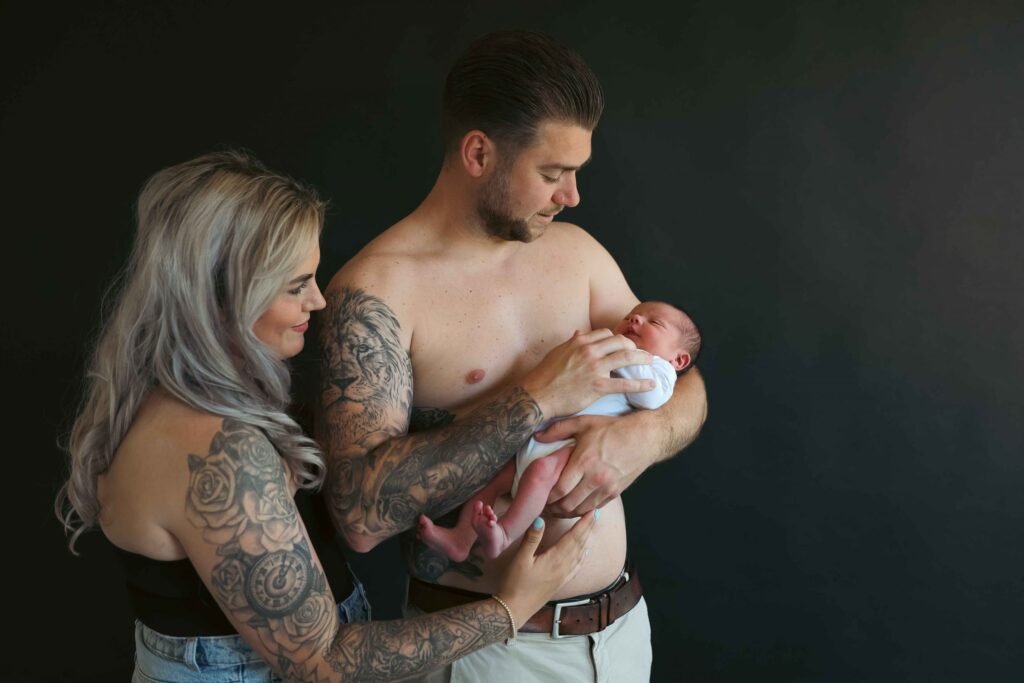 Tattooed parents holding their newborn in front of a dark backdrop during an intimate lifestyle portrait session.
