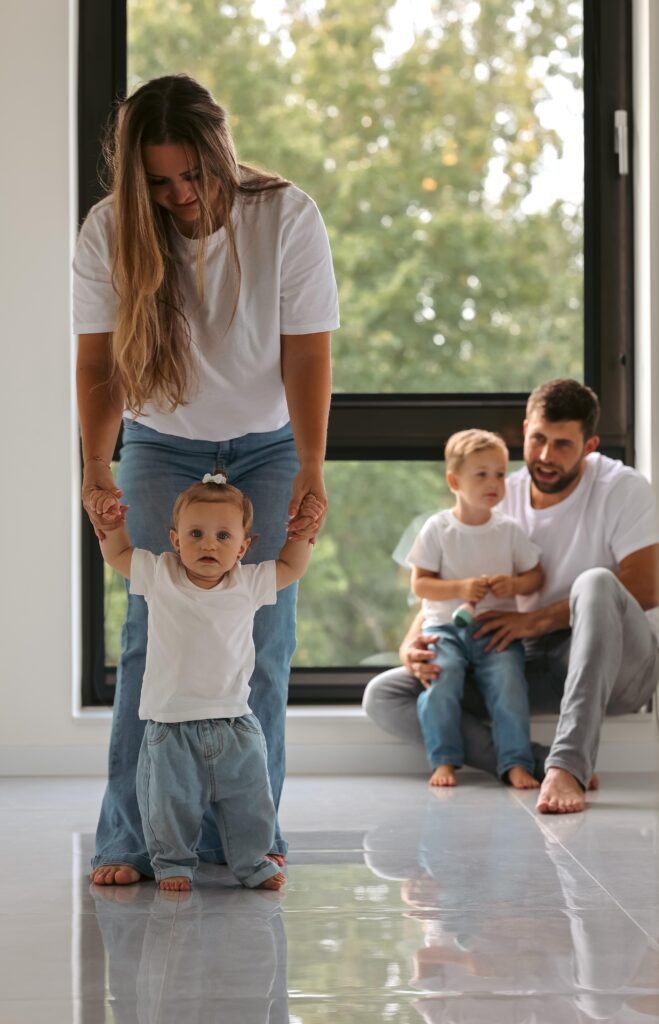 Toddler taking first steps with mother’s support while father and brother watch during a bright natural-light family session.