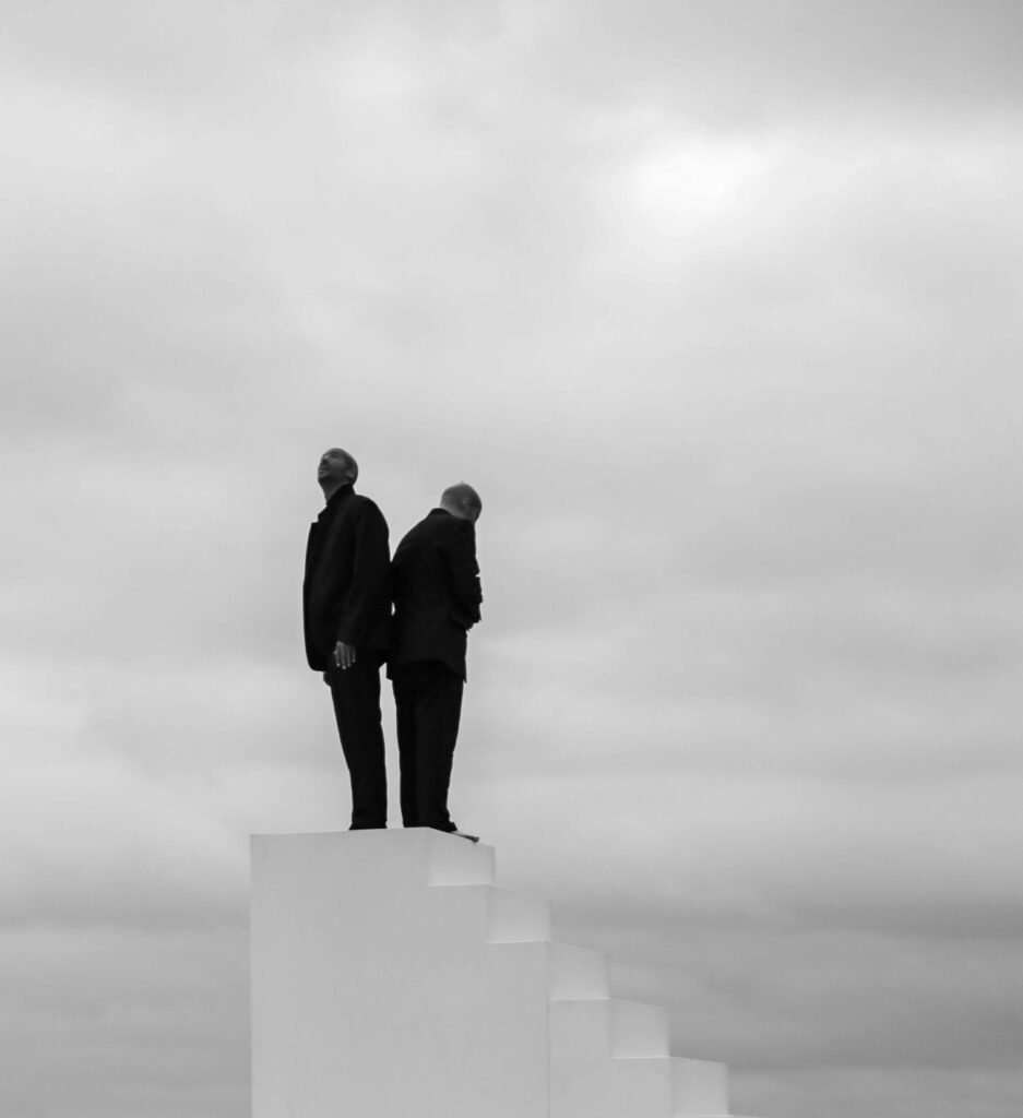 Two figures in dark clothing standing on a white staircase under a cloudy sky