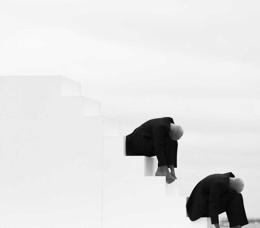 A surreal fine art photograph of two faceless figures in black suits positioned on stark white steps under a cloudy sky in The Hague.
