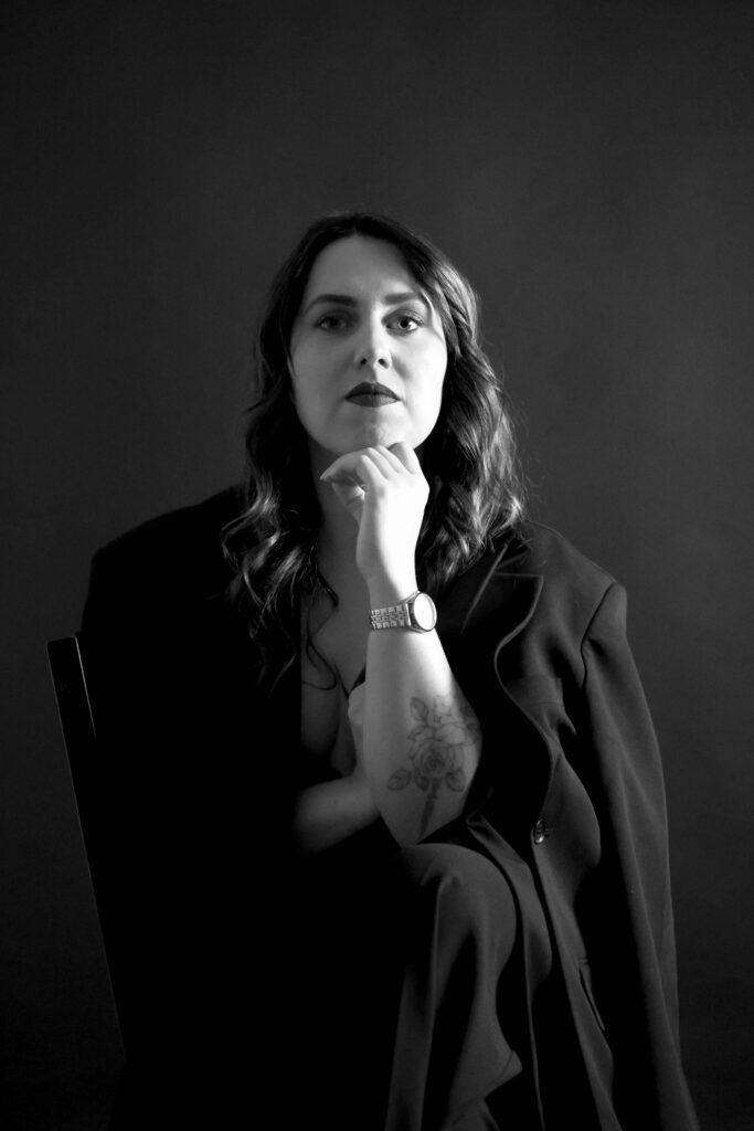 Black-and-white editorial portrait of a woman seated with hand under chin in a dark studio – The Hague.