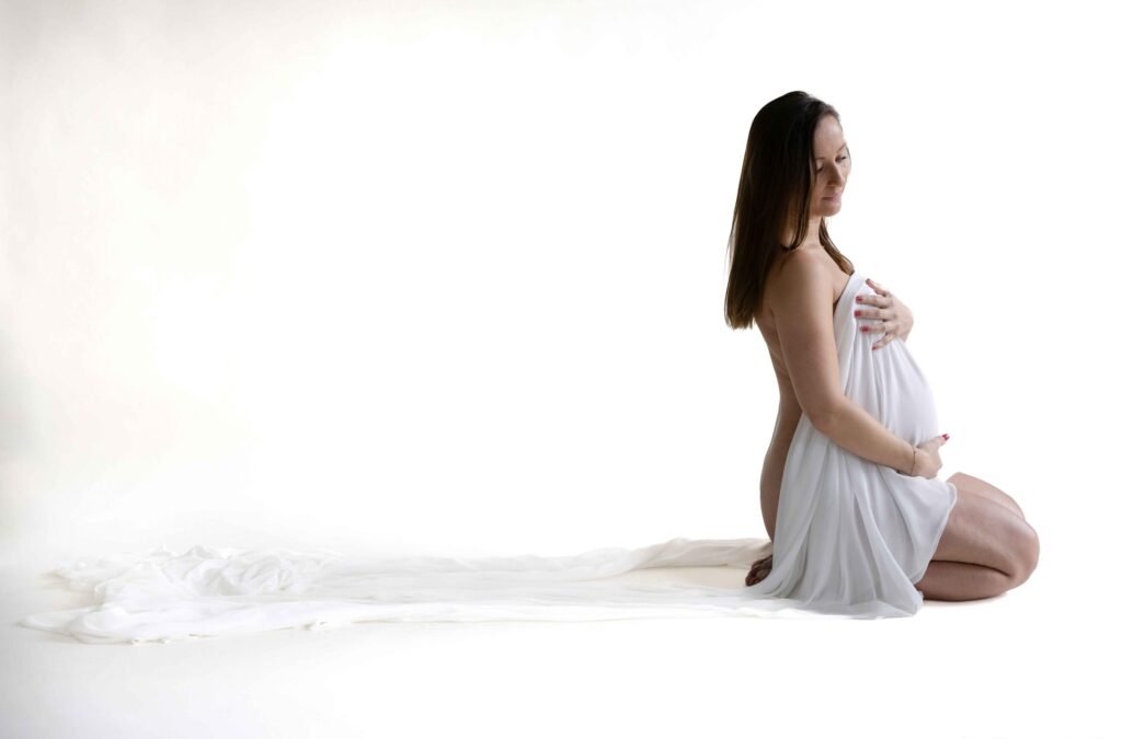 Pregnant woman kneeling and draped in white fabric in a bright, minimalist studio in The Hague.