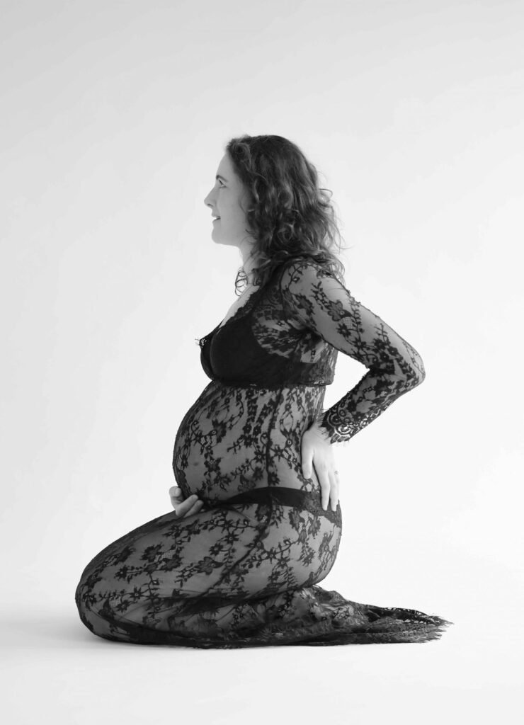 Profile view of a pregnant woman in a sheer black lace gown holding her belly in a studio in The Hague.
