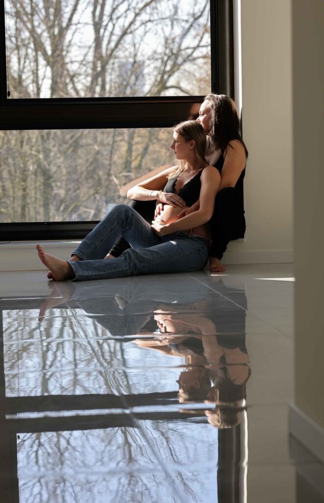 Couple sitting together with pregnant belly framed in window light and floor reflection in The Hague.