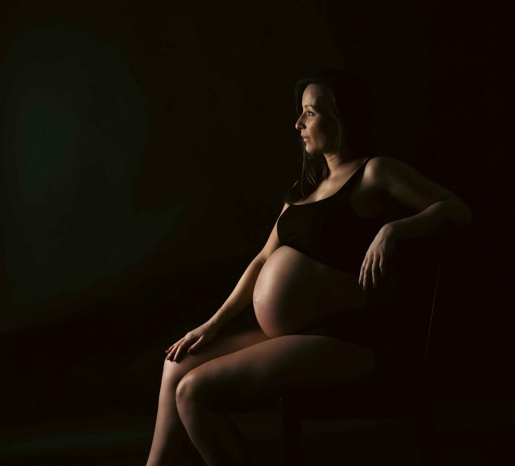 Pregnancy portrait in dramatic low light with expecting mother seated against a dark background in The Hague.