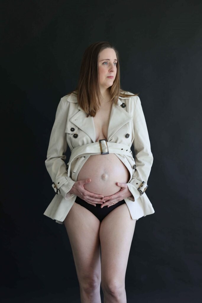 Pregnant woman wearing an open trenchcoat in studio for a maternity session in The Hague.