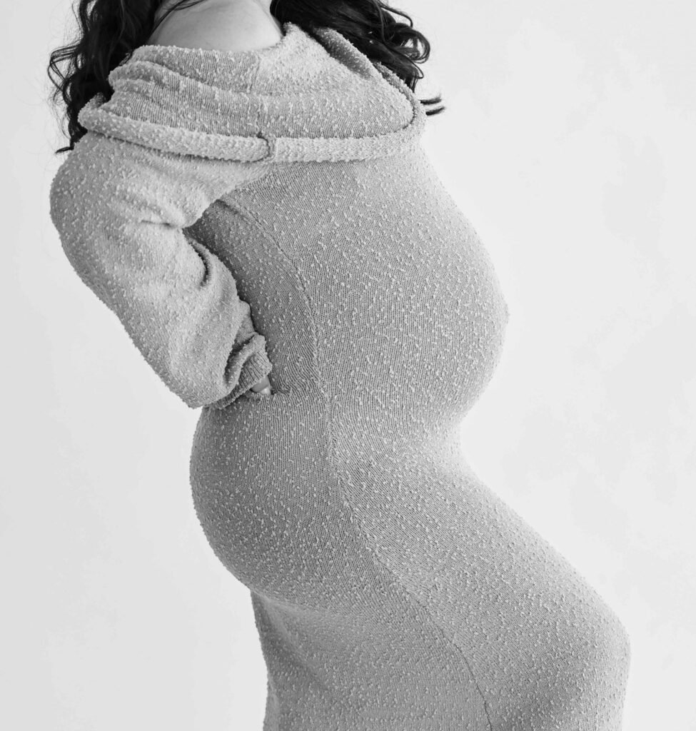 Close-up side view of a pregnant woman in a textured knit dress holding her lower back during a maternity shoot in The Hague.