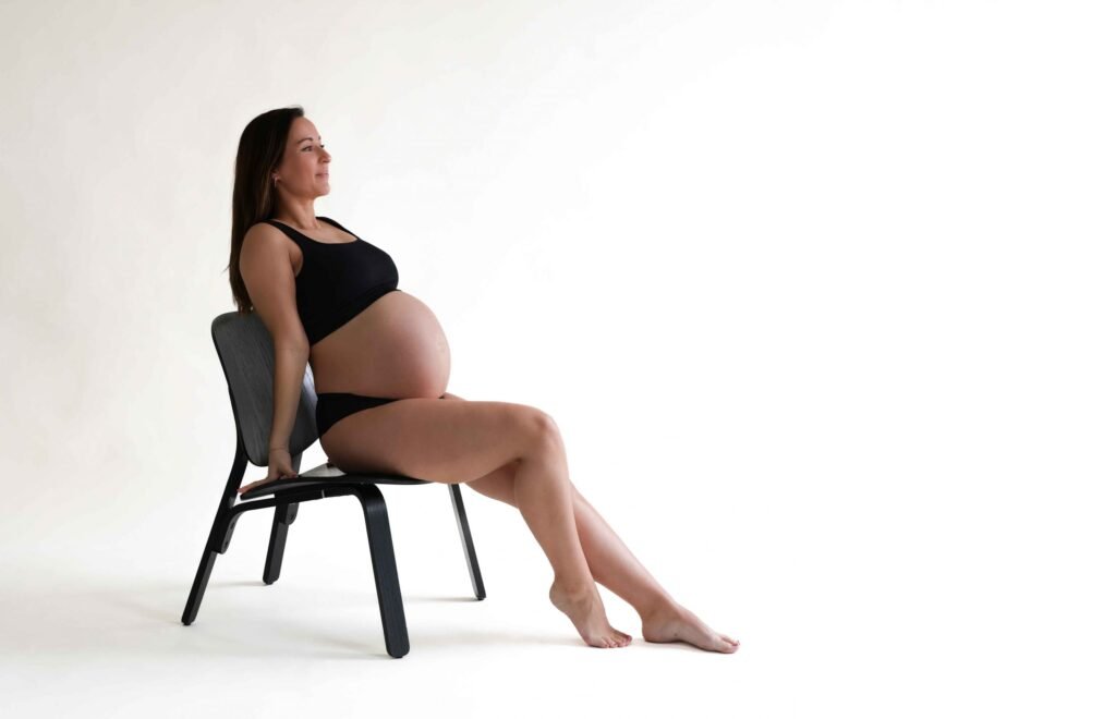 Pregnant woman sitting on a chair in a minimalist white studio holding her belly during a maternity shoot in The Hague.