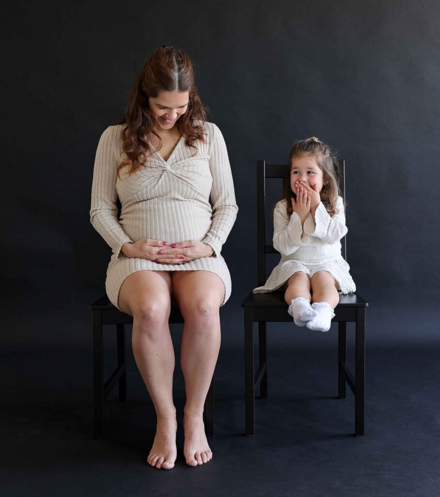 Pregnant mother sitting beside her young daughter who is smiling and covering her mouth during a maternity photoshoot.