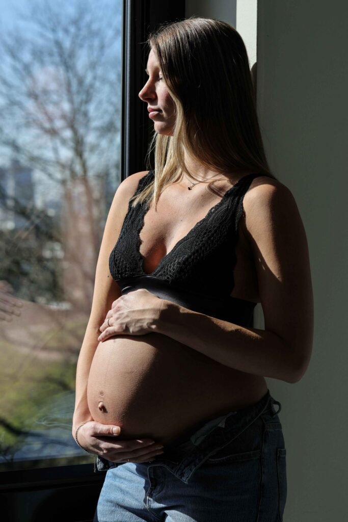 Pregnant woman standing by a window with natural light, hands on her bare belly during a maternity shoot in The Hague.