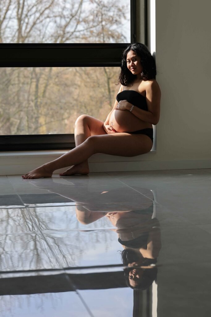Maternity portrait of expecting mother sitting by the window with soft reflection on the floor in The Hague.”